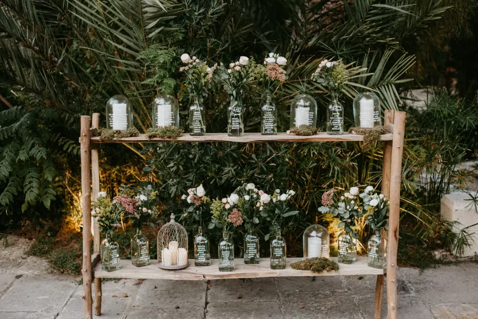 Estantería de madera de dos niveles con frascos de vidrio y velas dispuestas entre plantas y flores al aire libre.