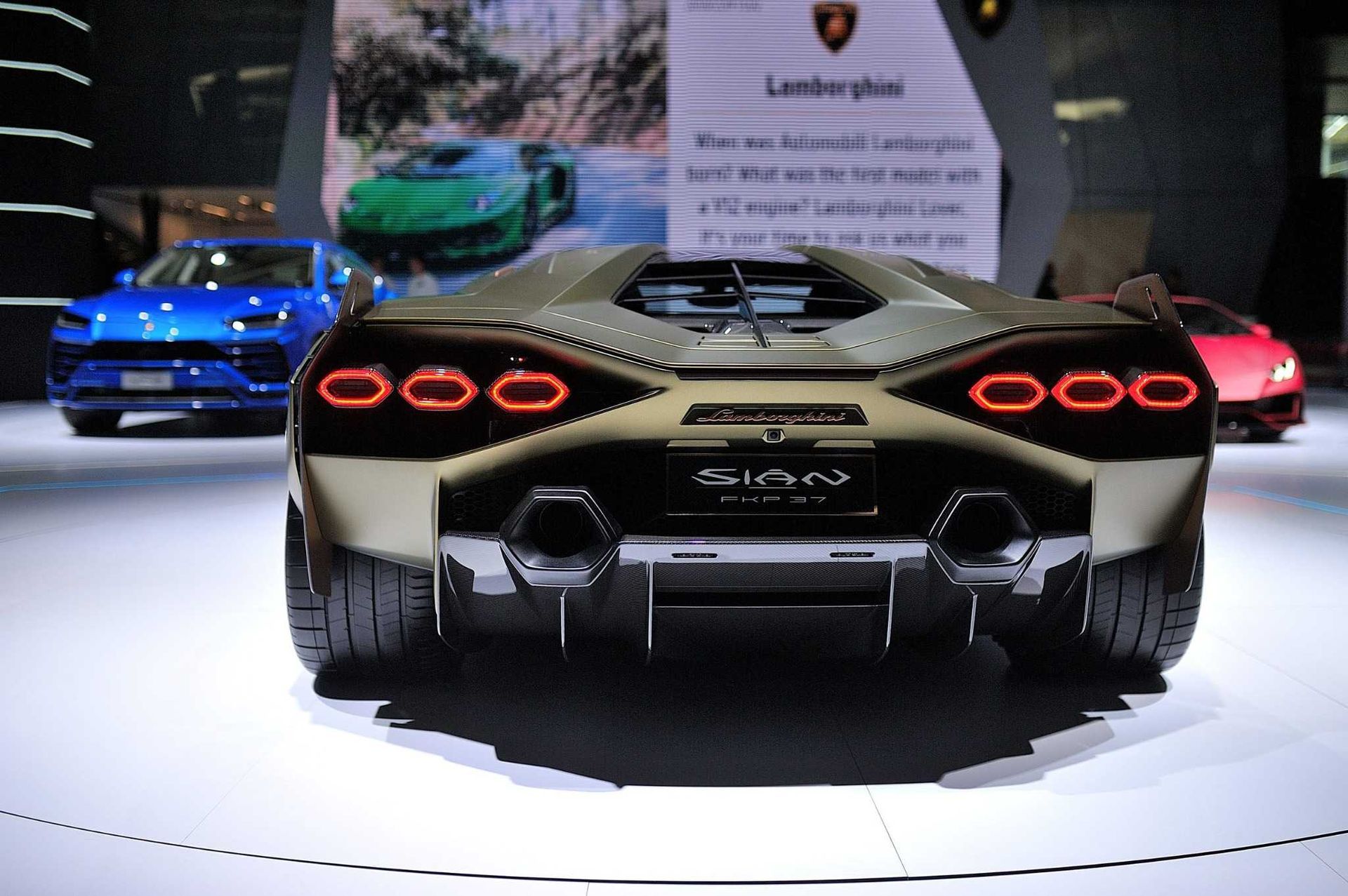 Vista trasera de un deportivo Lamborghini dorado en exhibición, con autos azules y rojos al fondo.