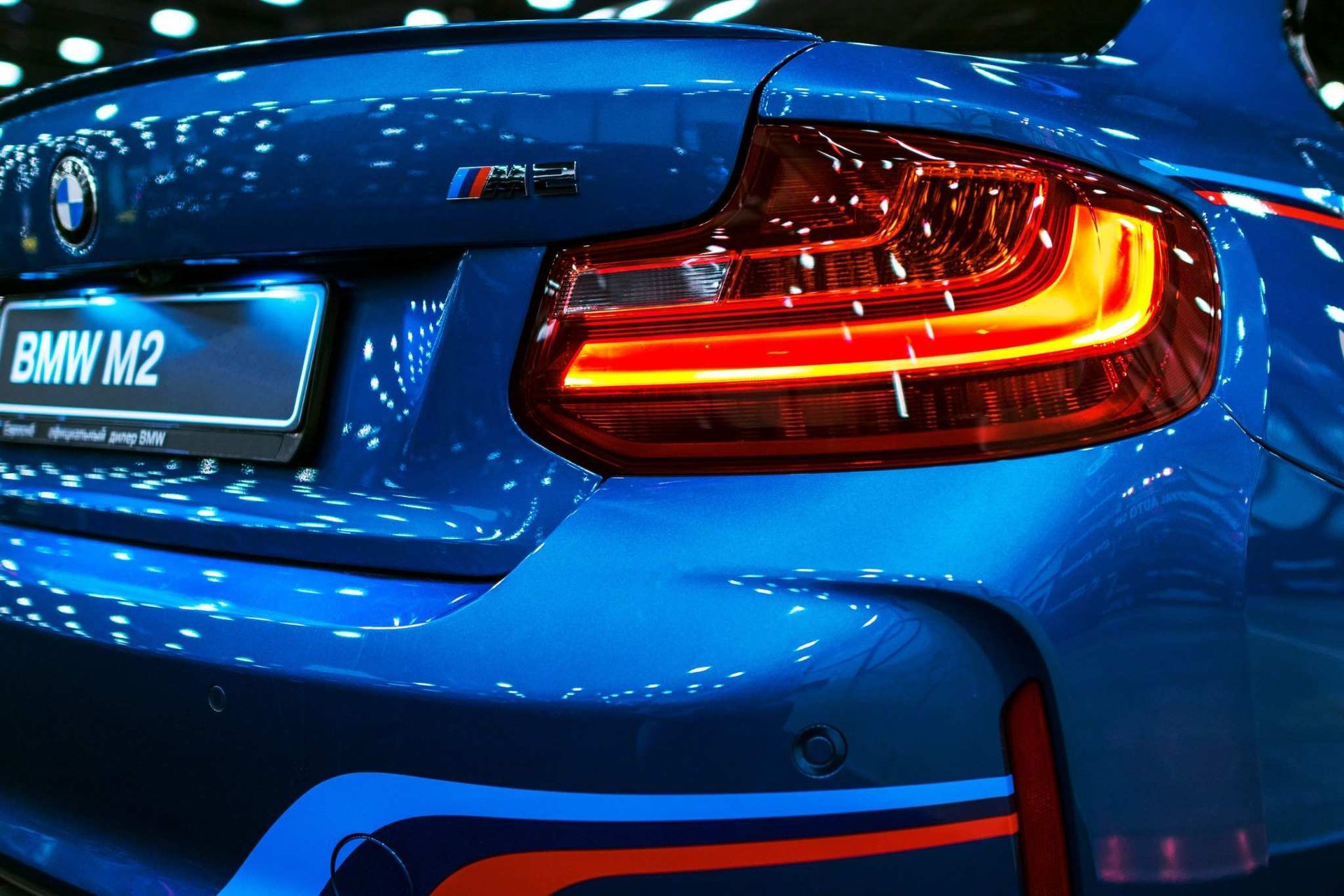Parte trasera de un BMW M2 azul con luces traseras y matrícula iluminadas en una exposición de automóviles.