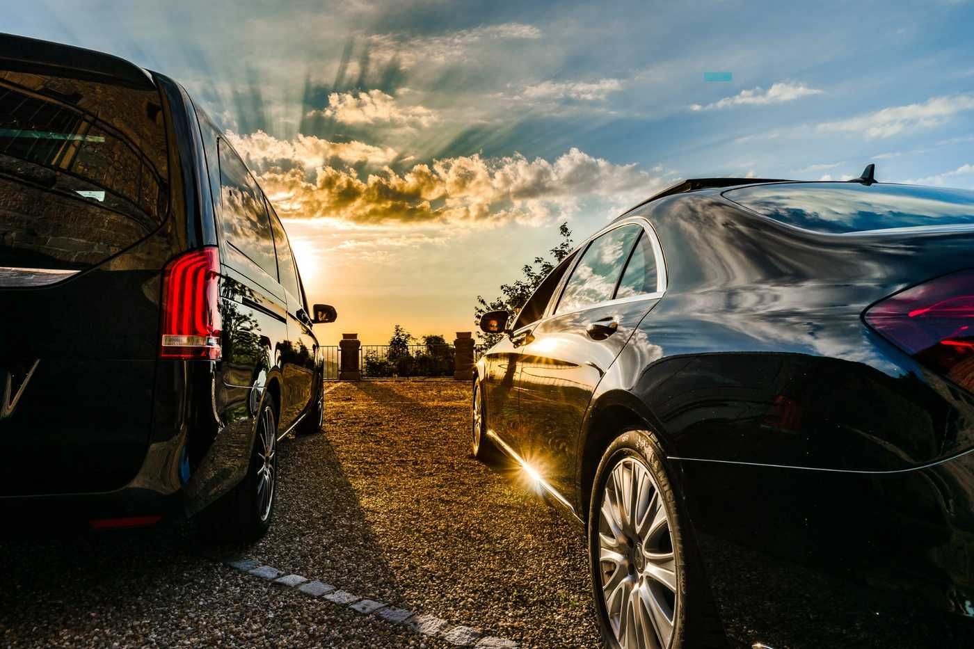 Autos de lujo estacionados al atardecer en un camino de grava, con luz dorada y nubes en el cielo.