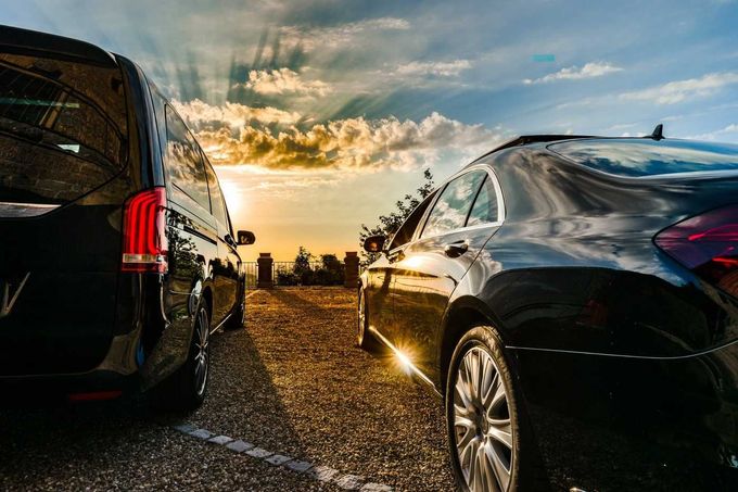 Autos de lujo estacionados al atardecer en un camino de grava, con luz dorada y nubes en el cielo.