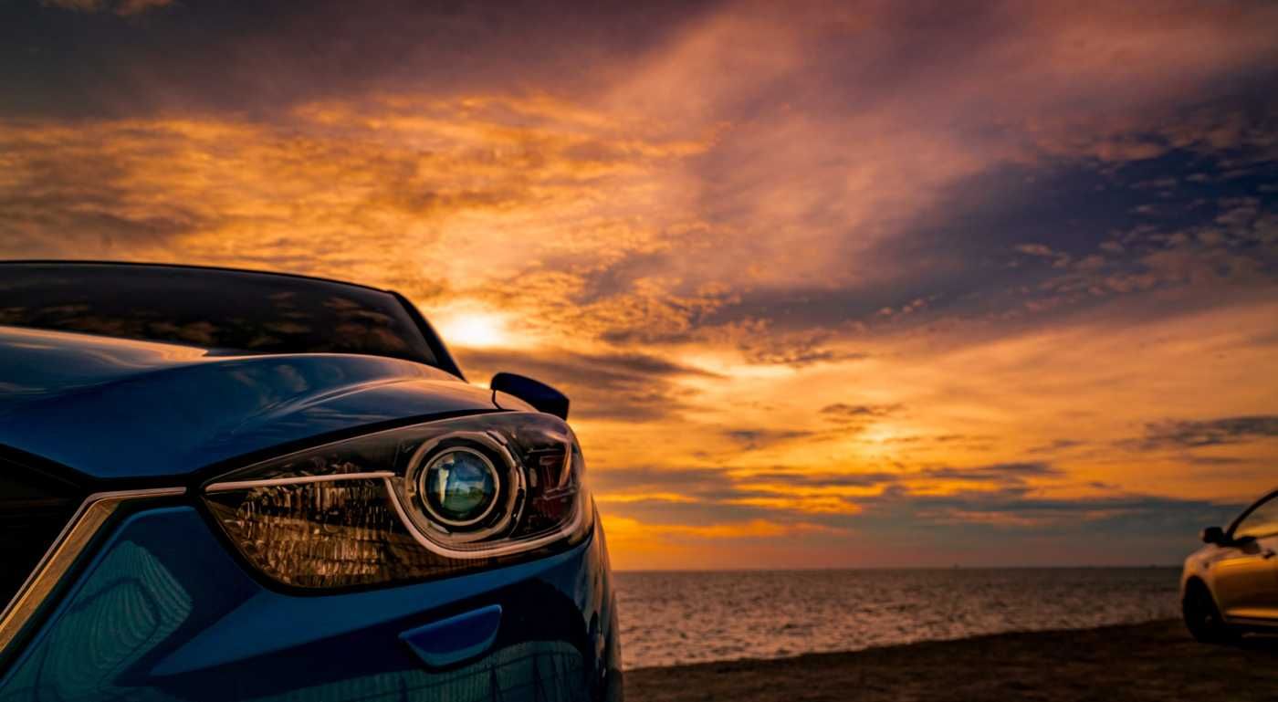 Un coche azul al atardecer junto al océano, con espectaculares nubes naranjas y un segundo coche a la derecha.