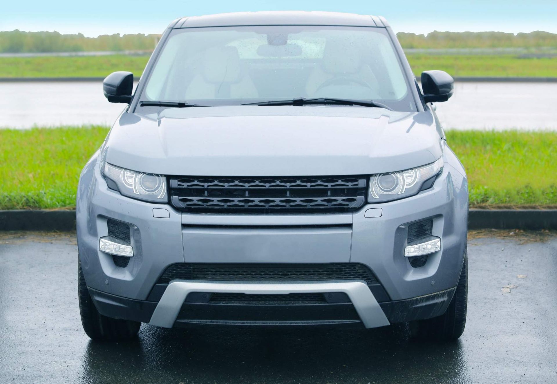 Vista frontal de un SUV gris estacionado en una carretera mojada junto a un campo verde.