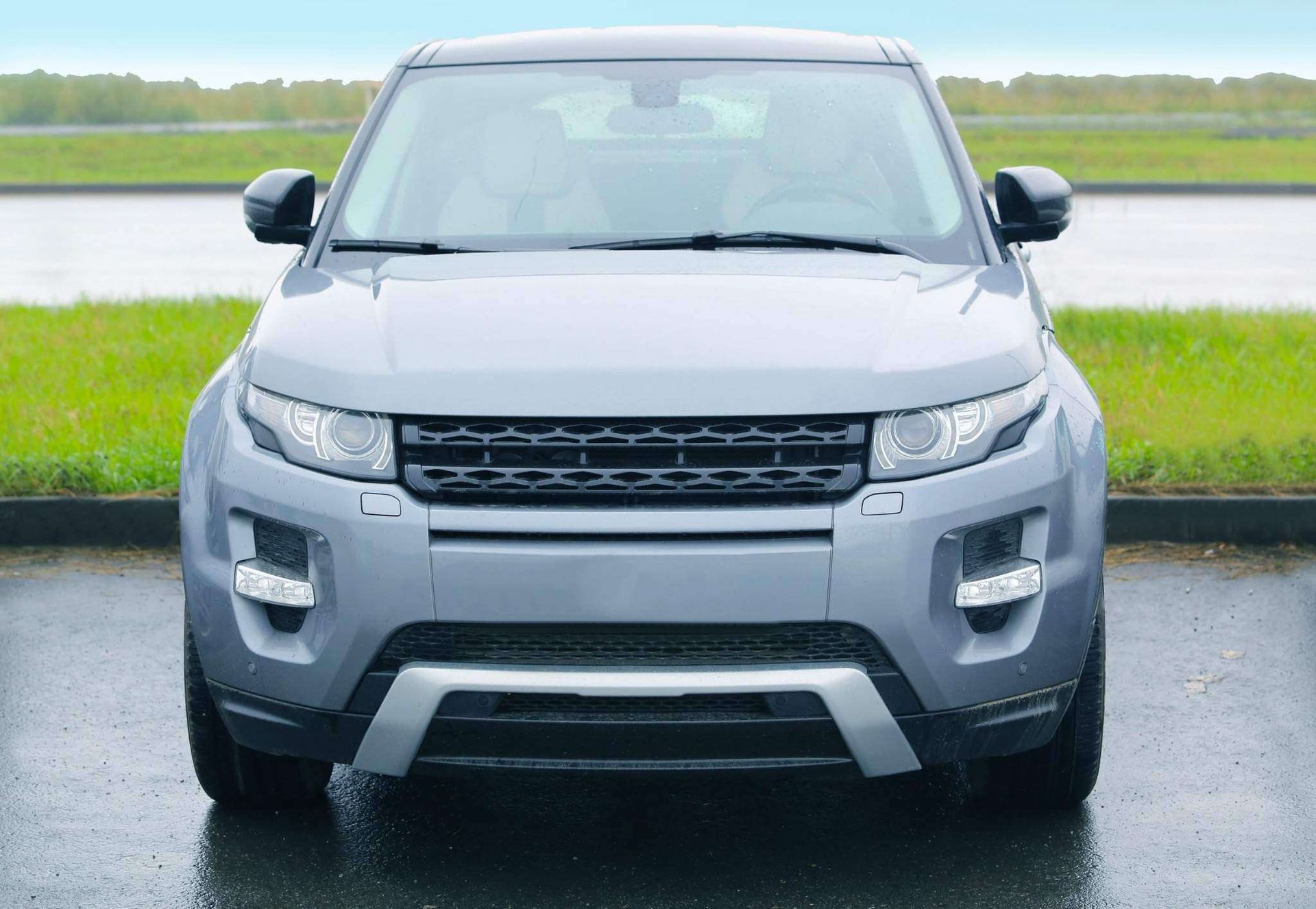 Vista frontal de un SUV gris estacionado en una carretera mojada junto a un campo verde.