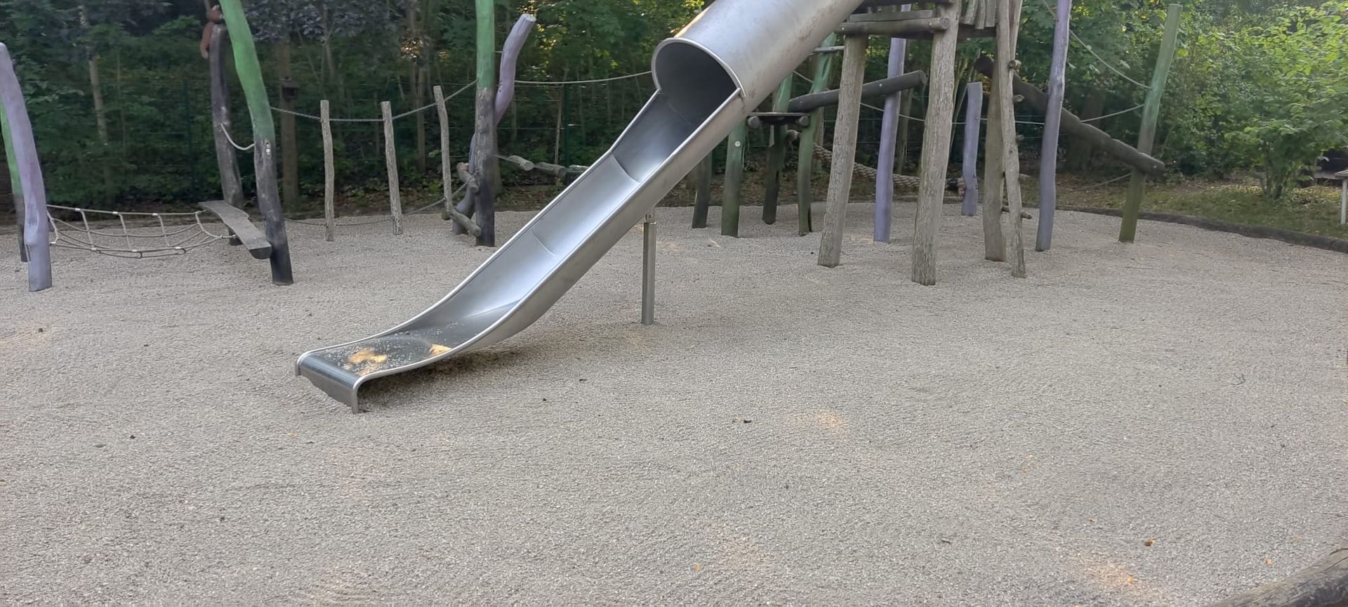 Eine Metallrutsche auf einem Spielplatz, umgeben von kleinen Kieselsteinen, Holzkonstruktionen und Bäumen.