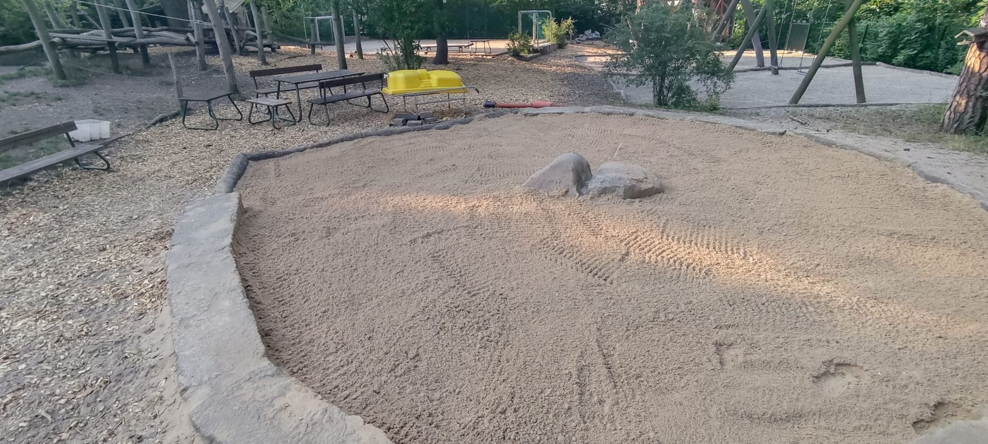 Ein Sandkasten mit einem Stein in der Mitte, umgeben von einem Betonweg und Bäumen.