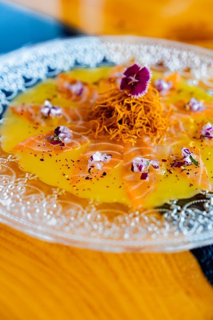 Plato de cristal transparente con postre amarillo decorado con hilos naranjas y una flor morada sobre una mesa de madera.