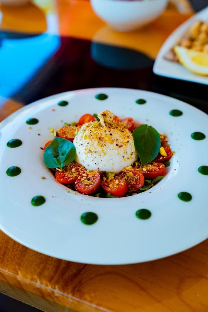 Plato gourmet con tomates cherry, burrata y albahaca en un plato blanco con puntos de salsa verde.