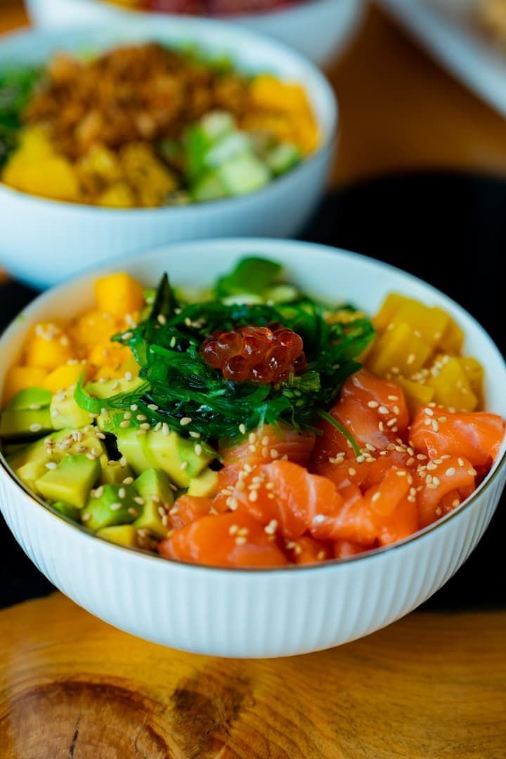 Tres coloridos poke bowls con salmón, aguacate y mango sobre una mesa de madera.