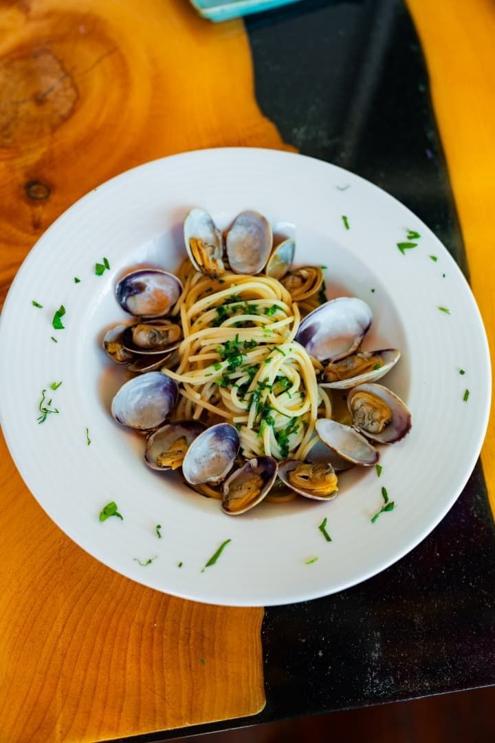 Almejas con pasta en un tazón blanco, adornadas con perejil sobre una mesa de madera.