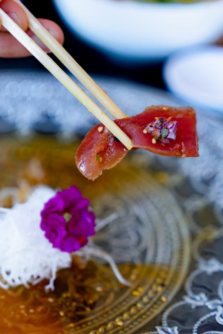 Palillos sosteniendo un bocado de sushi rojo brillante sobre un plato ornamentado con guarnición blanca y flor morada.