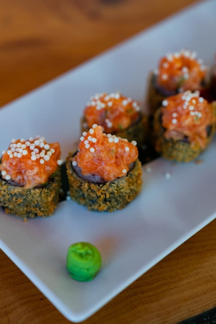 Rollos de sushi cubiertos con salmón y semillas de sésamo en un plato blanco, con wasabi aparte.