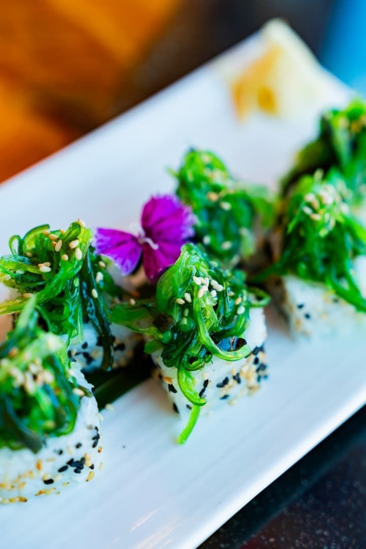 Rollitos de sushi cubiertos con guarnición verde sobre un plato blanco, decorados con una flor morada.