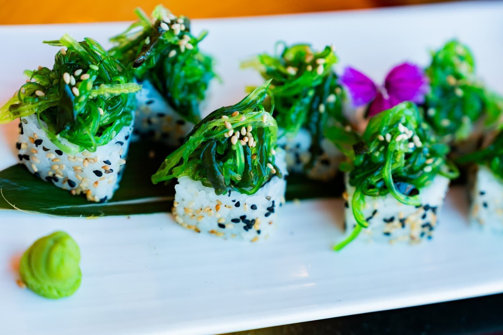 Rollitos de sushi cubiertos con verduras en un plato blanco, con wasabi y guarnición morada.