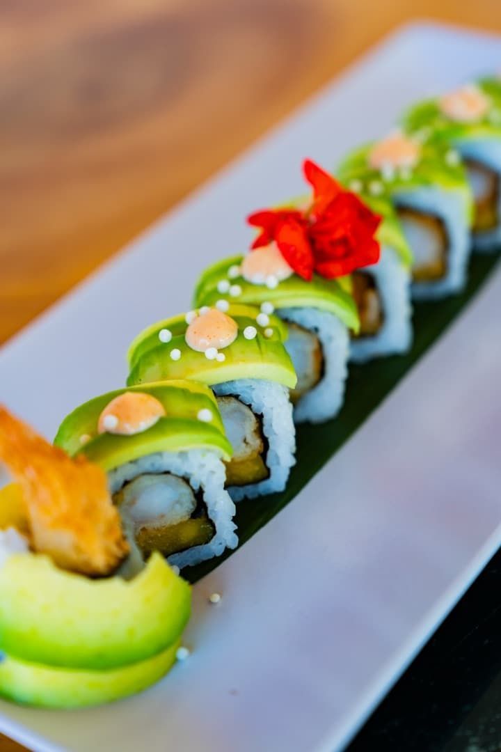 Rollos de sushi cubiertos con aguacate y salsa en un plato blanco, adornados con una flor roja.