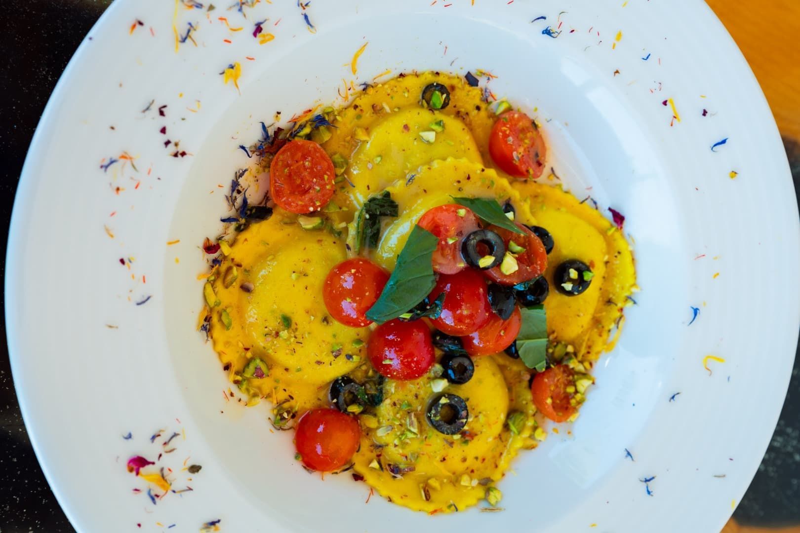 Pasta colorida servida en un plato con tomates cherry, aceitunas y hierbas aromáticas sobre un plato blanco.