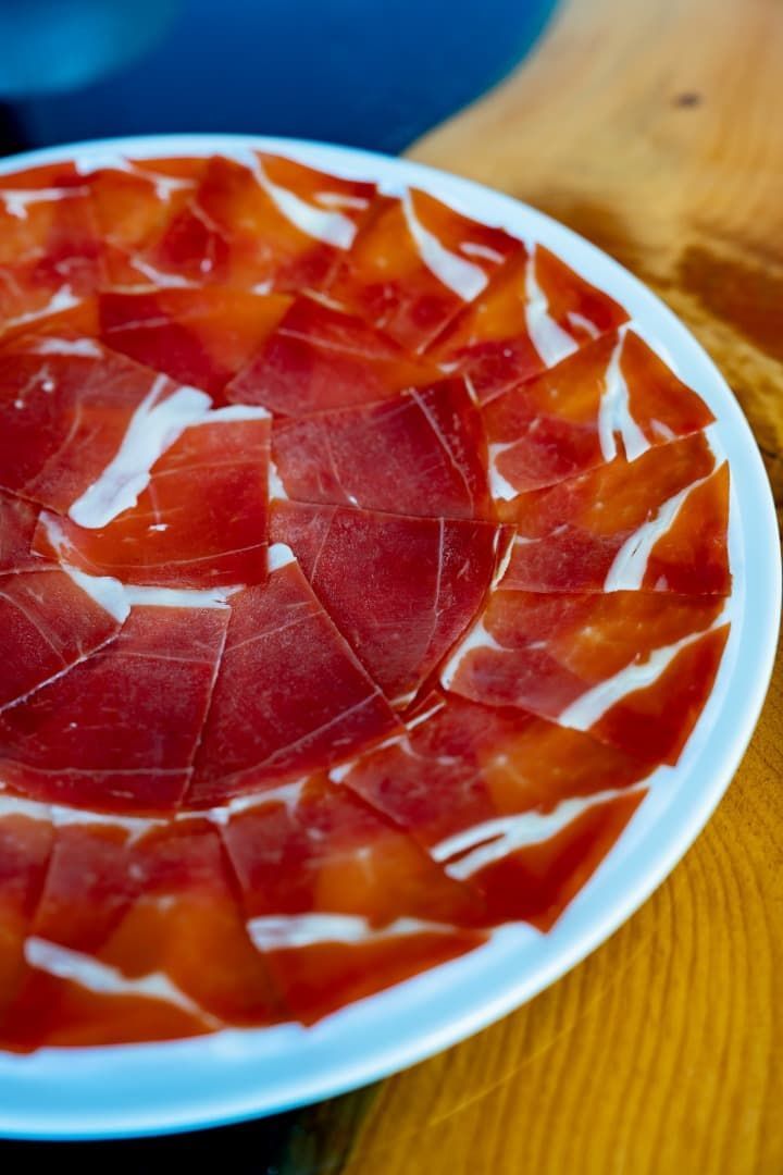 Jamón curado cortado en lonchas finas dispuesto en un plato blanco sobre una mesa de madera.