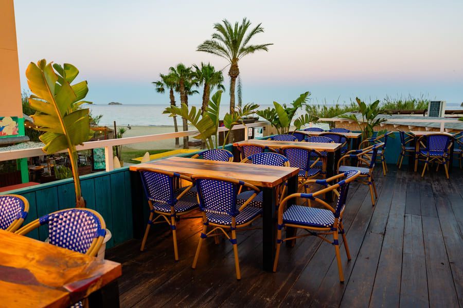 Terraza de restaurante al aire libre junto al mar con sillas azules, mesas de madera y palmeras al atardecer.