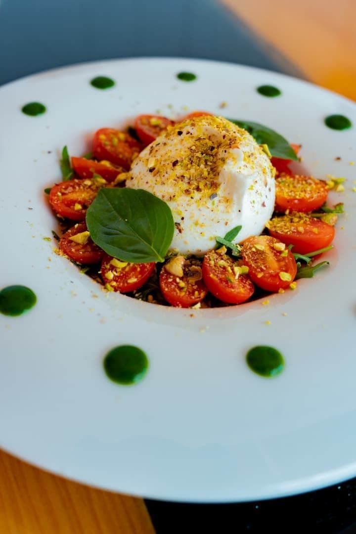 Ensalada Caprese con burrata, tomates cherry, albahaca y puntos de salsa verde sobre un plato blanco.
