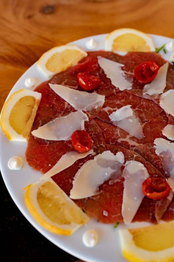 Plato de prosciutto con virutas de parmesano, fresas y rodajas de limón en un plato blanco.