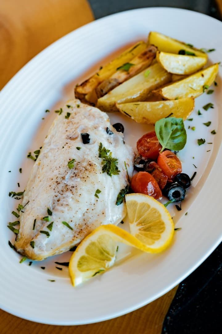 Pescado al horno con rodajas de limón, patatas asadas y tomates cherry en un plato blanco.