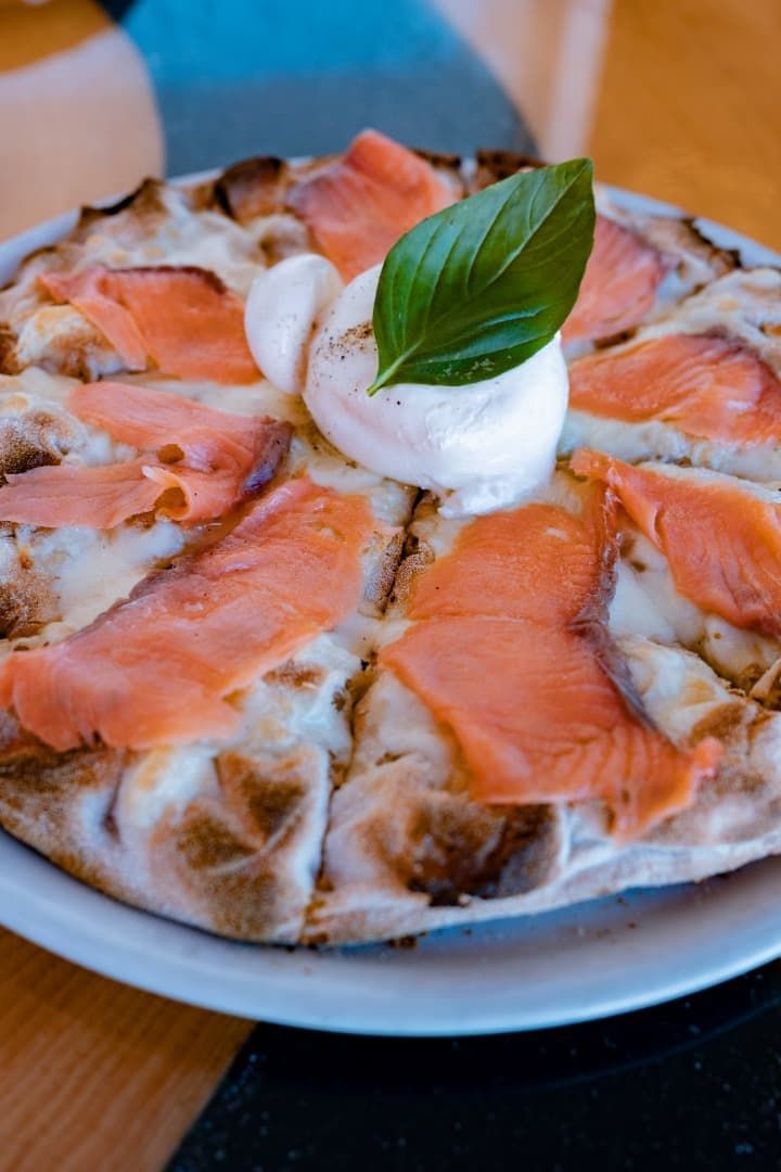 Pizza cubierta con rodajas de salmón, queso blanco y una hoja de albahaca en un plato.