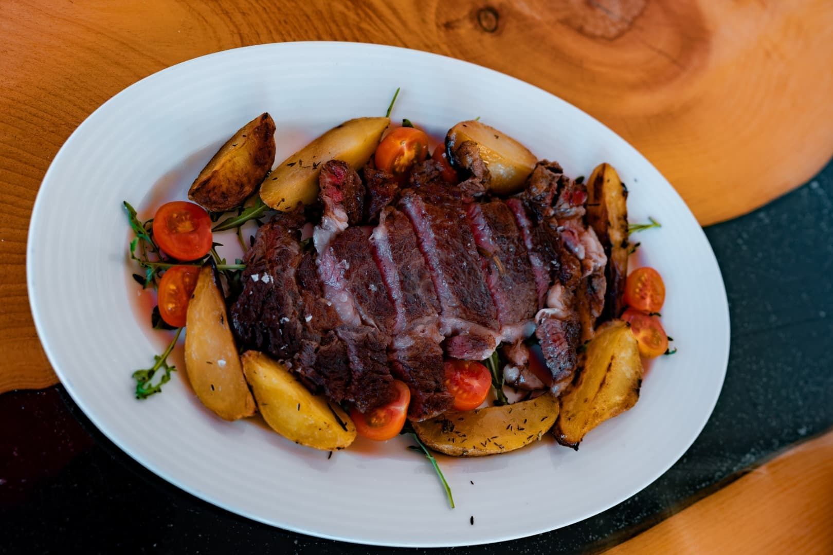 Filete en rodajas servido con patatas y zanahorias asadas en un plato blanco sobre una mesa de madera.