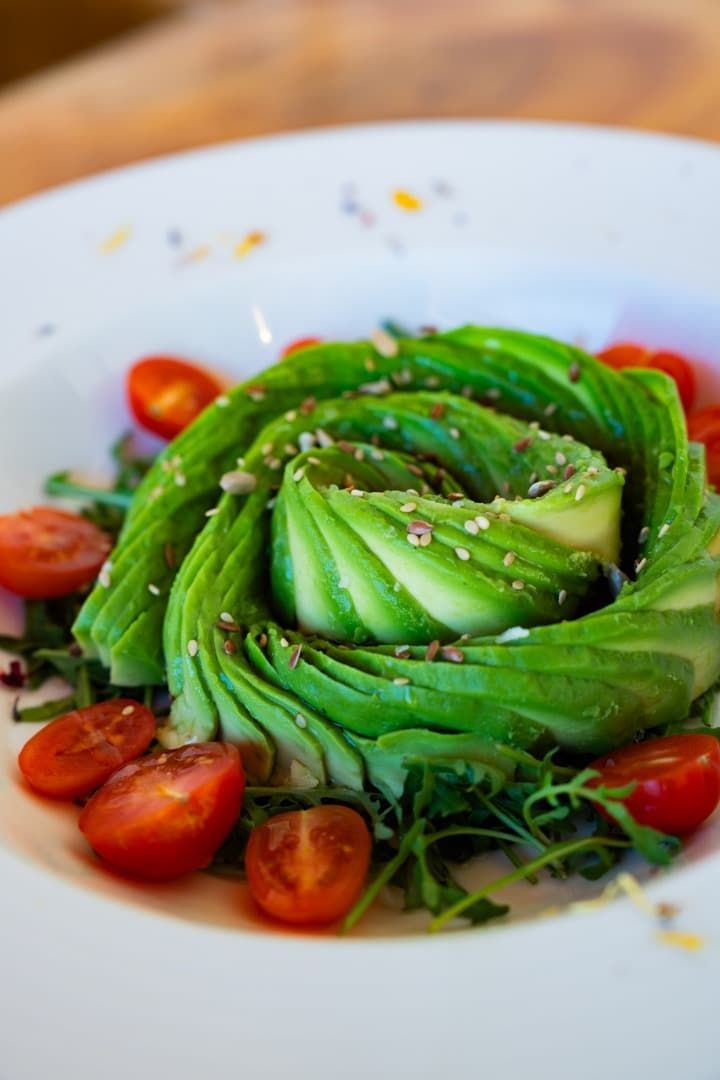 Ensalada verde en capas dispuesta como una rosa, cubierta con semillas y rodeada de tomates cherry en un plato blanco.