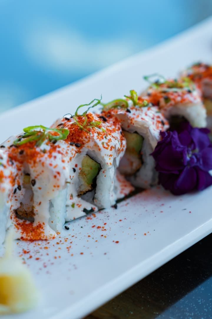 Rollitos de sushi rociados con salsa blanca y condimento rojo en un plato blanco, adornados con col morada.