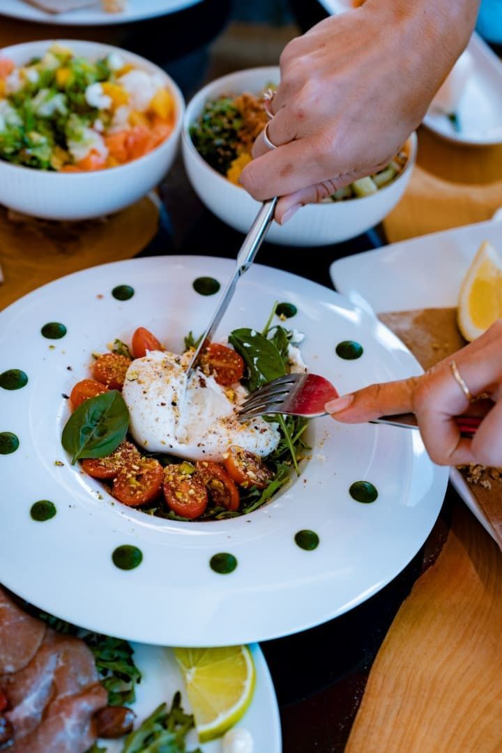 Manos emplatando una ensalada gourmet con queso, tomates y salsa en un plato blanco en la mesa de un restaurante.