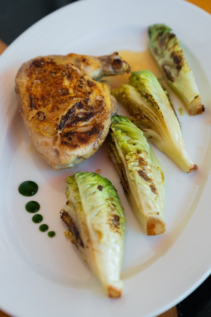 Pechuga de pollo a la parrilla con gajos de endivia chamuscada en un plato blanco, adornada con puntos de salsa verde.