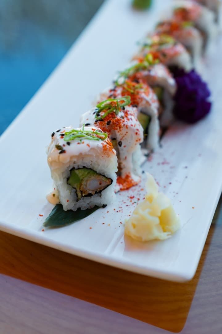 Rollo de sushi cubierto con salsa y hierbas en un plato rectangular blanco con jengibre y wasabi.