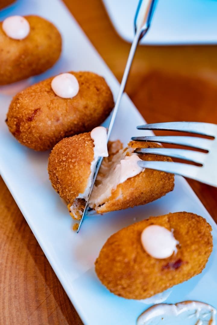 Croquetas empanadas con salsa blanca en un plato, una de ellas abierta con tenedor y cuchillo.