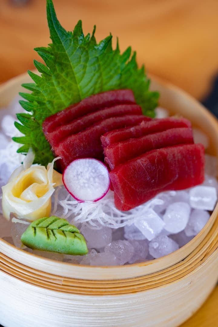 Sashimi de atún en rodajas sobre hielo en una cesta de bambú, adornado con hoja de shiso, daikon y rodaja de limón.