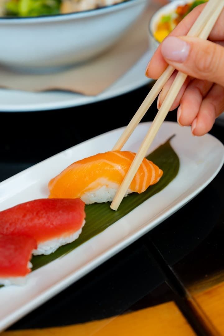 Una mano usa palillos para tomar sushi de un plato blanco con guarnición verde.