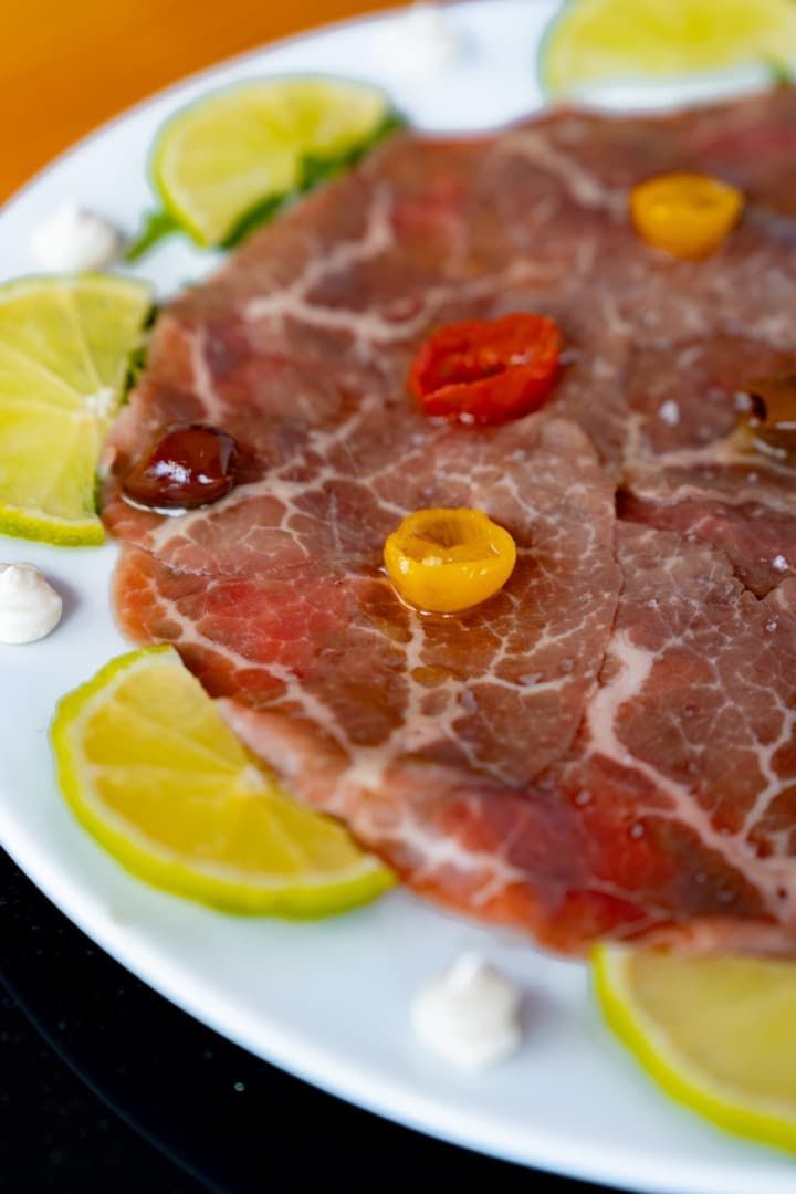 Carne curada cortada en finas lonchas, cubierta con tomates cherry en un plato, adornada con rodajas de lima y pequeñas cucharadas de queso.