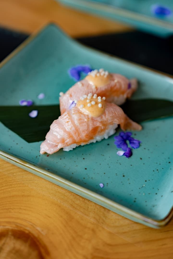 Dos piezas de sushi de salmón en un plato color turquesa con pétalos de flores moradas.