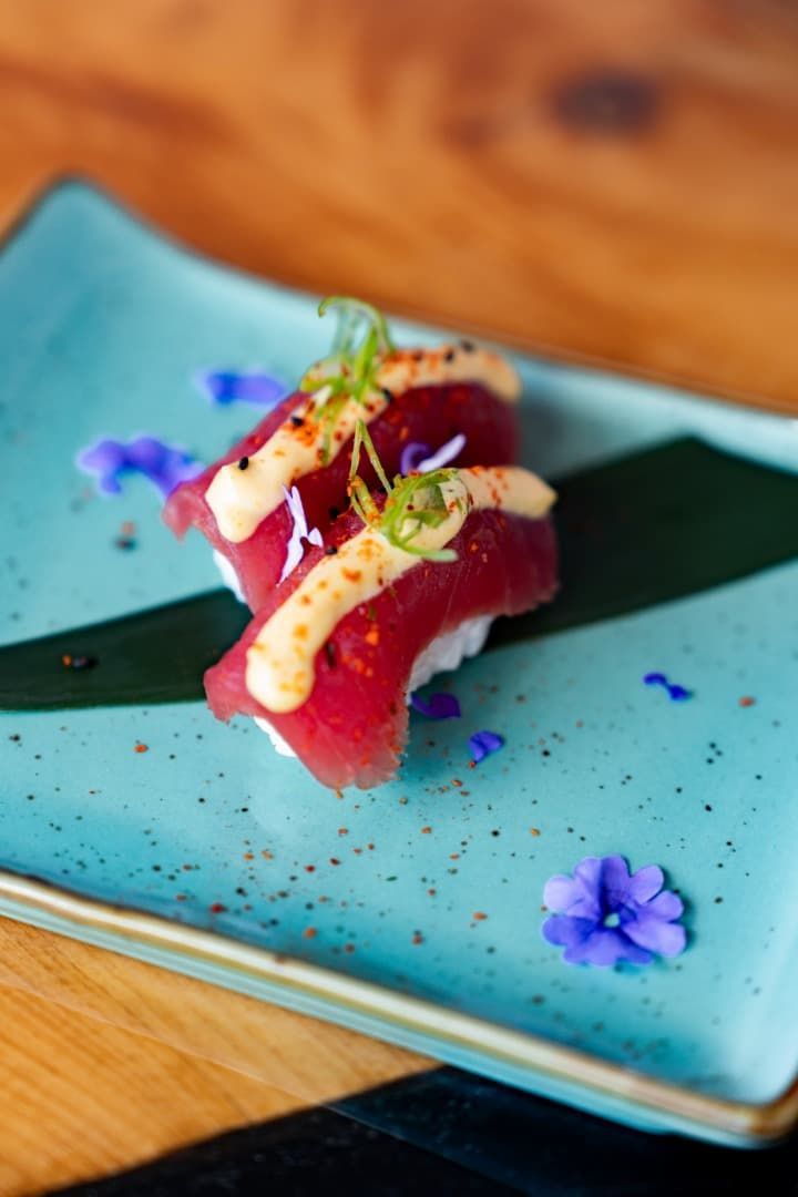 Sushi gourmet emplatado en un plato rectangular azul con adornos de flores moradas.