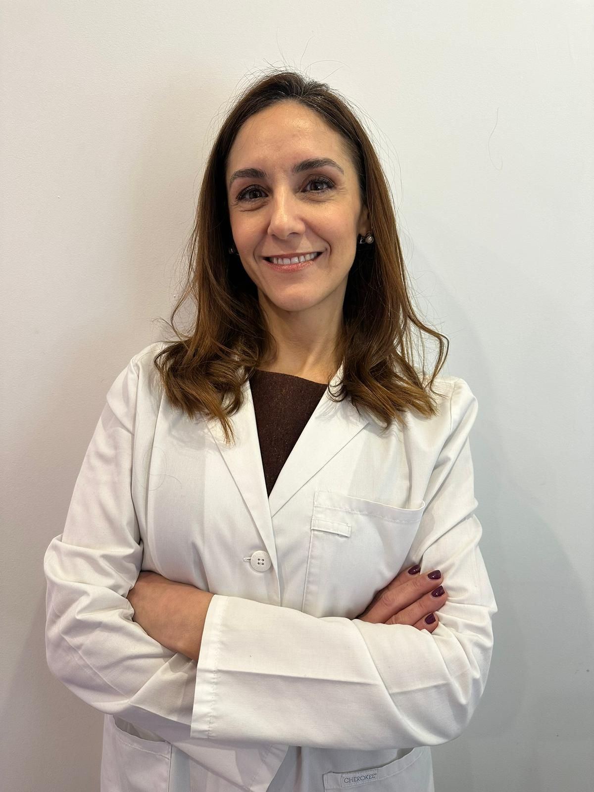 Mujer con bata blanca de laboratorio y brazos cruzados, sonriendo a la cámara.