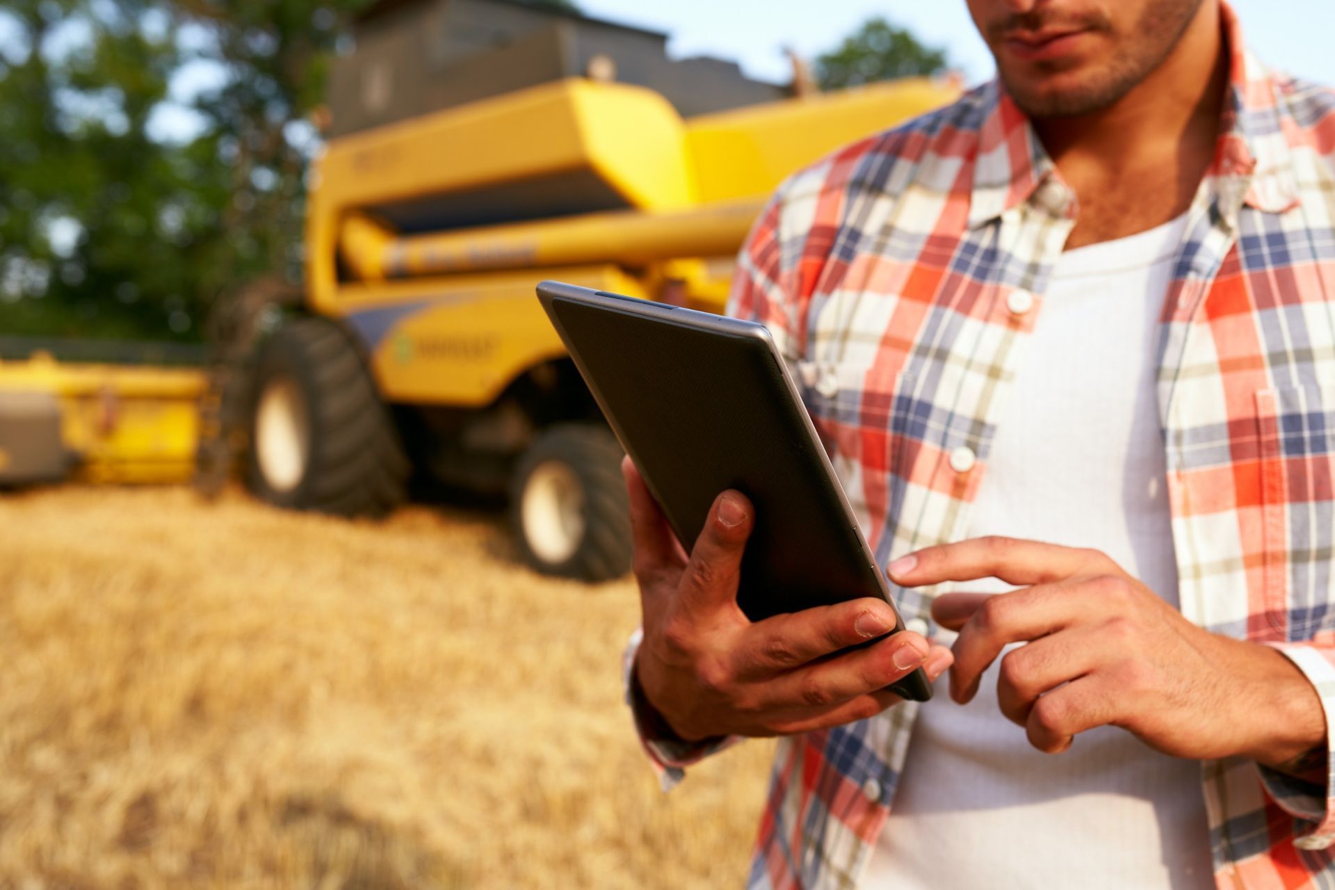 Un hombre está usando una tableta en un campo con un tractor al fondo.