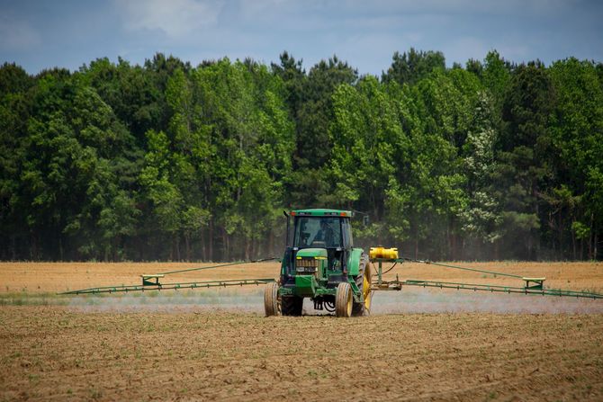 Un tractor verde está rociando un campo con árboles al fondo.
