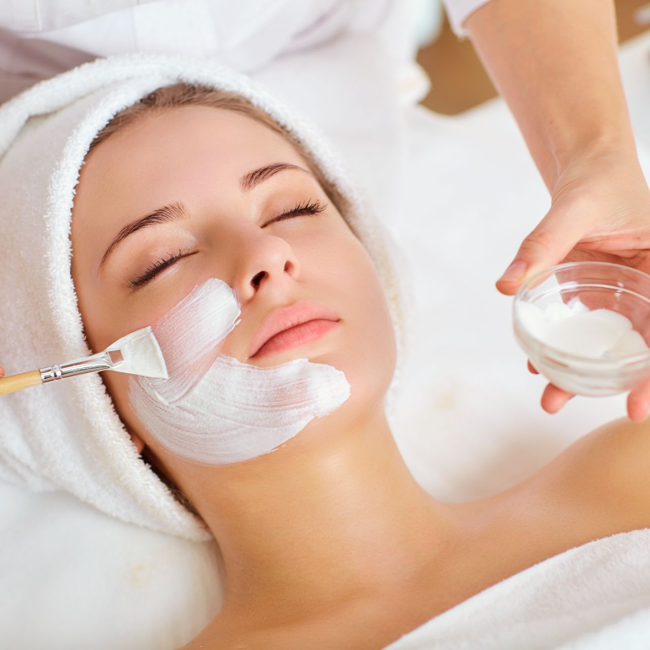 Masque blanc en train d'être appliqué sur le visage d'une femme qui a une serviette sur la tête