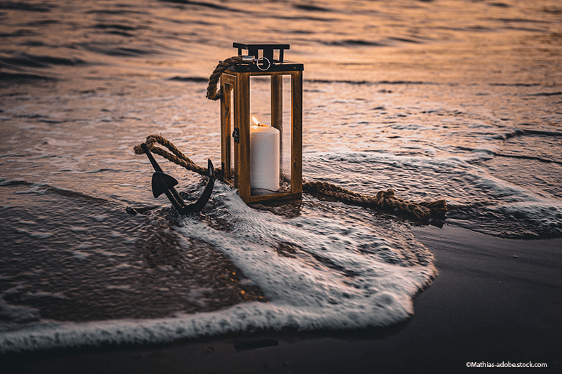 Laterne mit brennender Kerze und Anker an einem Strand mit einlaufenden Wellen bei Sonnenuntergang.