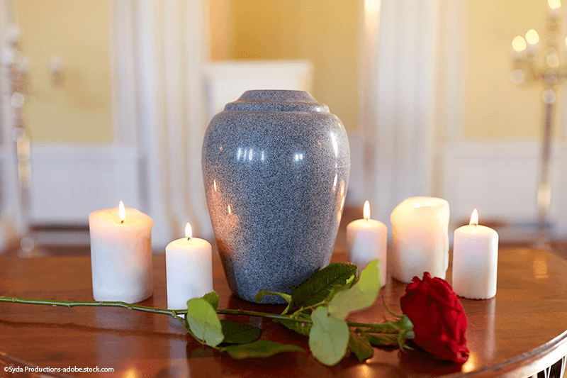 Eine grau gesprenkelte Urne, umgeben von brennenden Kerzen und einer roten Rose auf einem Holztisch.