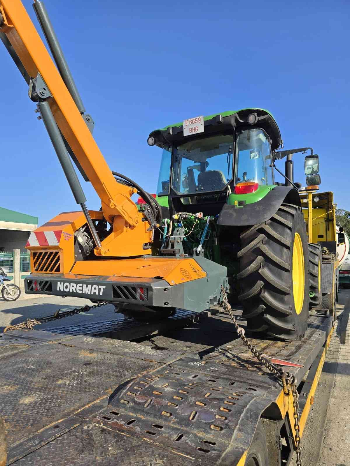 Una grúa naranja carga un tractor verde en un remolque de plataforma bajo un cielo azul despejado.