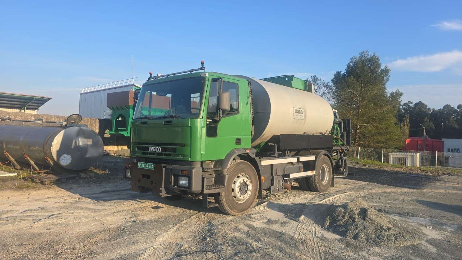 Camión hormigonera verde estacionado en un terreno de grava bajo un cielo despejado.