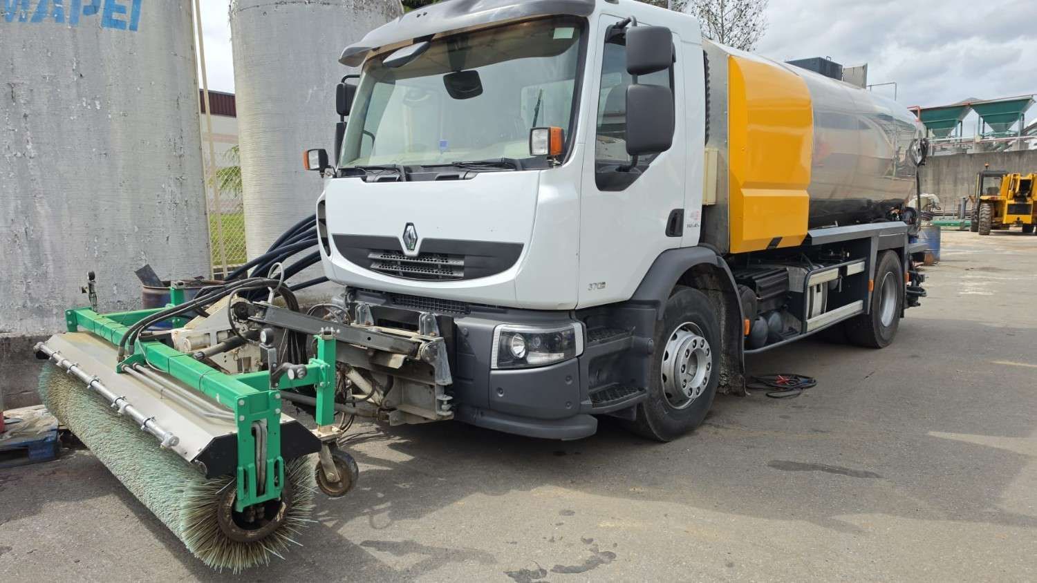 Una excavadora amarilla está estacionada junto a un tractor verde frente a un edificio.