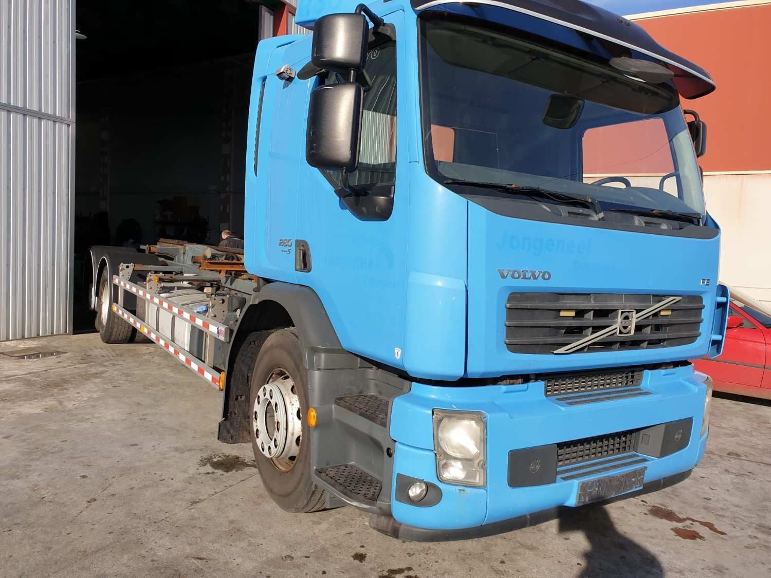 Camión Volvo azul de plataforma plana estacionado frente a la entrada de un almacén.