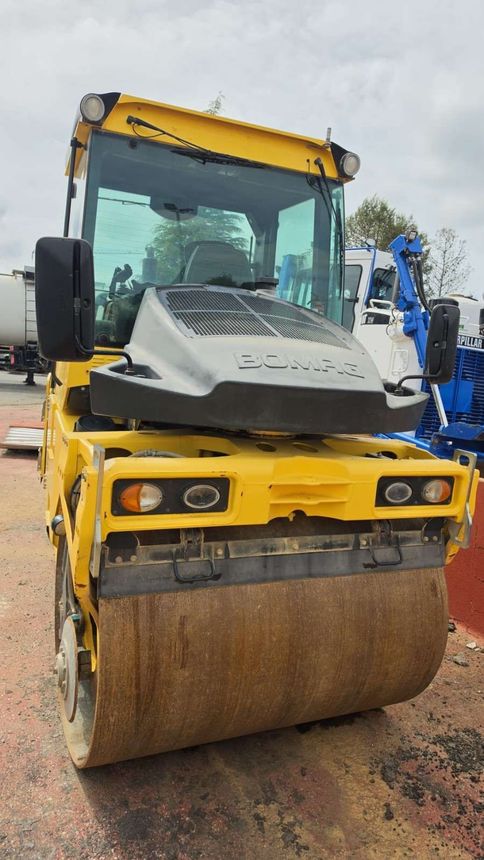 Una excavadora amarilla está estacionada junto a un tractor verde frente a un edificio.
