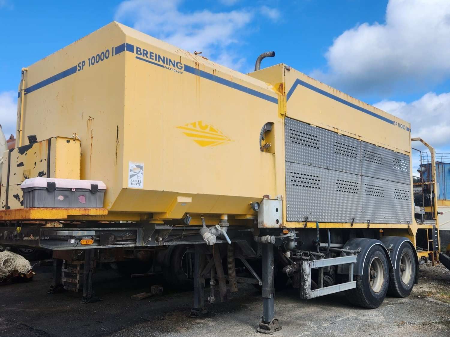 Máquina móvil industrial amarilla sobre un remolque, con rejillas de ventilación y patas de apoyo, estacionada al aire libre bajo un cielo azul.
